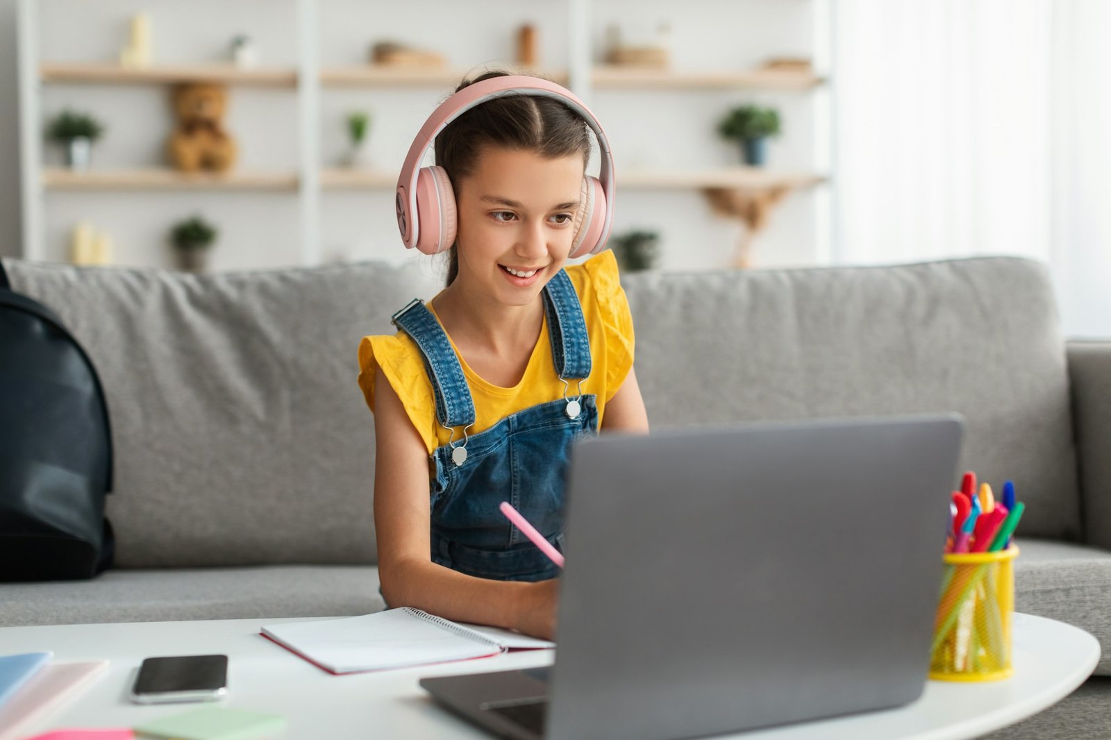 Schoolgirl in wireless headset sitting at desk, writing in textbook